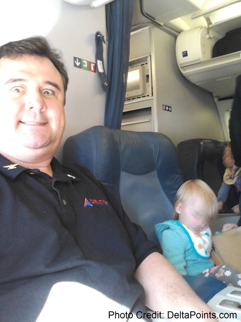 A man is taking a selfie on an airplane. He is wearing a black polo shirt with a logo. Next to him, a young child is asleep in the seat, wearing a blue hoodie with a colorful design. The airplane interior, including seats and overhead compartments, is visible in the background.
