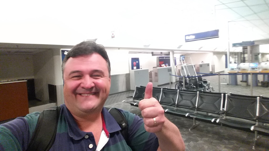 A person is taking a selfie in an airport terminal. They are smiling and giving a thumbs-up gesture. The background shows empty seating, a wheelchair, and some check-in counters. The area appears to be quiet and not crowded.