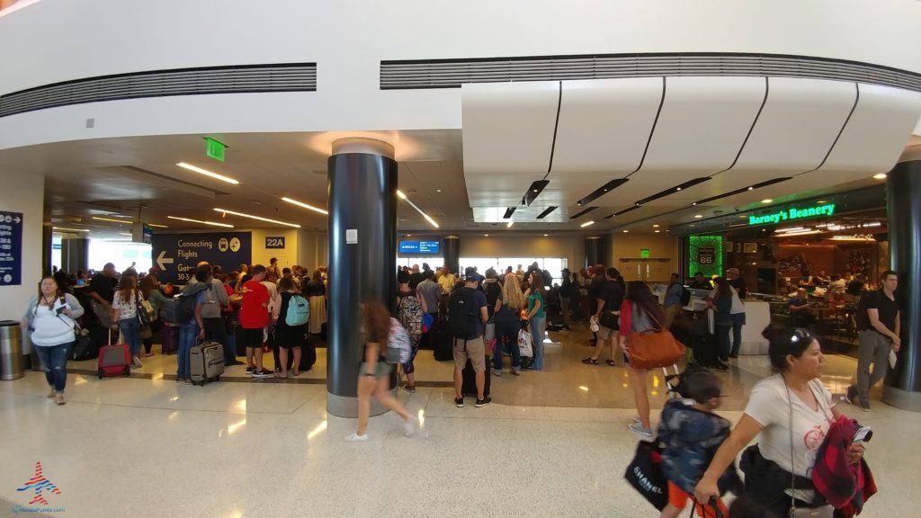 The image shows a busy airport terminal with a large group of people gathered near a gate area. There are signs indicating connecting flights and gate numbers. To the right, there is a restaurant called "Barney's Beanery." People are carrying luggage and some are walking through the terminal. The atmosphere is bustling and crowded.