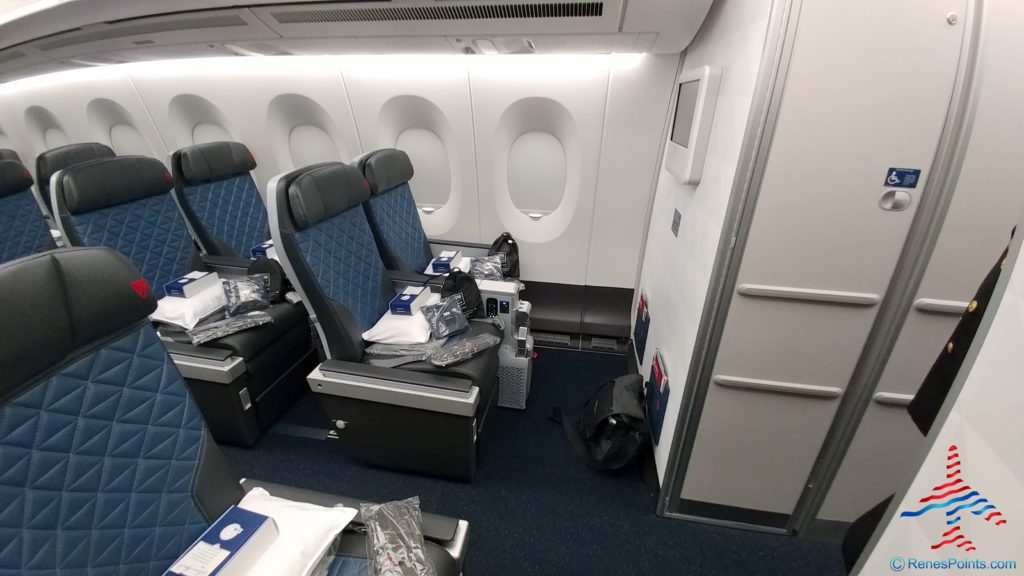 The image shows the interior of an airplane cabin, specifically a section with several rows of premium or business class seats. The seats are upholstered in blue with a diamond pattern and have headrests. Each seat has a pillow and a blanket placed on it, along with a small amenities kit. The cabin is well-lit, and there are windows along the side. The image also shows a bulkhead wall with a small screen and a sign indicating accessibility features.