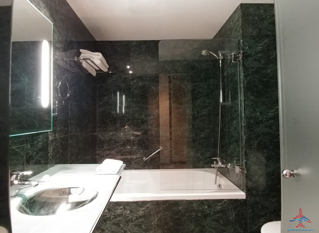 This image shows a modern bathroom with dark marble walls. There is a bathtub with a glass shower screen and a showerhead above it. A white towel is placed on the edge of the tub. To the left, there is a countertop with a sink and a large mirror with a vertical light strip. Towels are neatly stacked on a shelf above the bathtub.