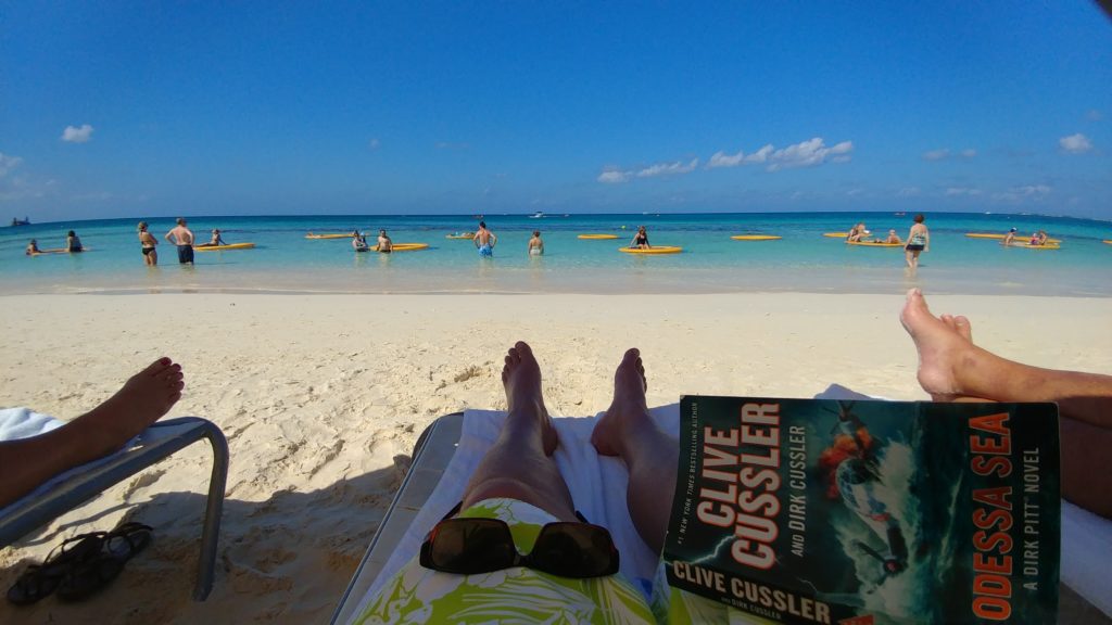 a person's legs on a beach with a book and people in the background