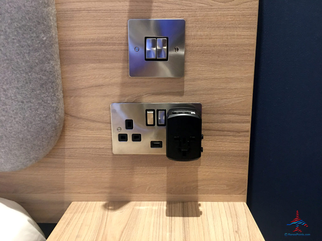 A nightstand, power outlets, and Gimars travel adapter are seen inside a room at the Holiday Inn Express London Heathrow T4 (LHR airport hotel) in Hounslow, United Kingdom.