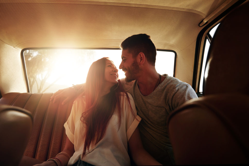 Happy couple on road trip. Young man and woman sitting on rear seat of car looking at each other smiling on a summer day.