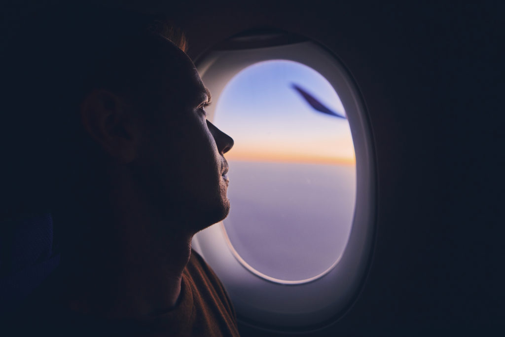 Silhouette of young man traveling by airplane. Pensive passenger looking through window during flight at sunrise.