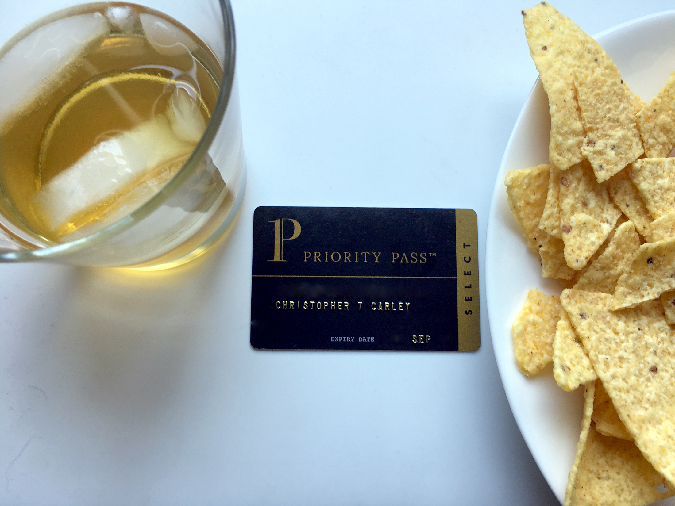 A Priority Pass Select membership card is seen between a Scotch on the rocks and plate of corn chips.