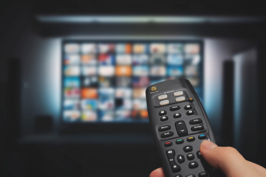 A person is holding a remote control in front of a large TV screen. The TV displays a blurred grid of colorful thumbnails, likely representing a streaming service or media library. The room is dimly lit, emphasizing the glow from the screen.