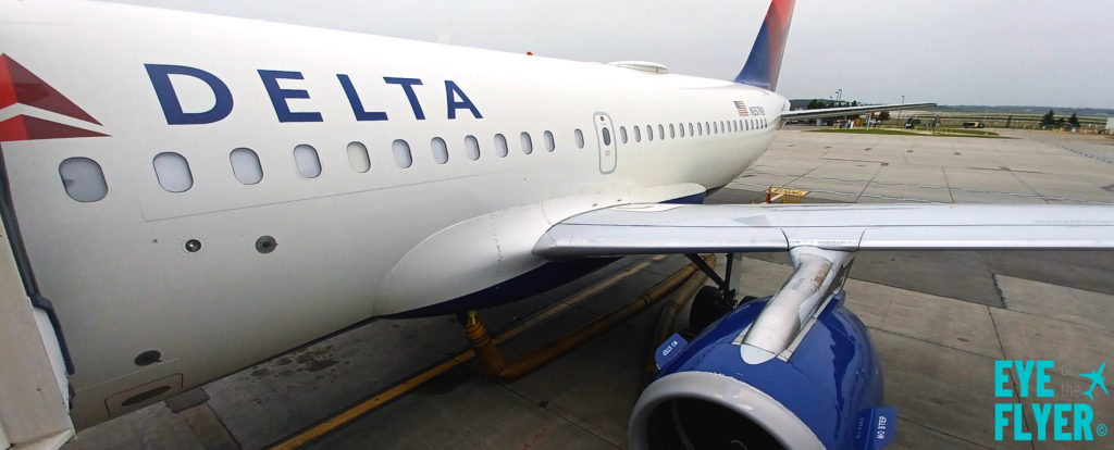 The image shows a Delta Air Lines aircraft parked on an airport tarmac. The view is from the side, showing the airline's logo and part of the fuselage, wing, and engine. The background includes the airport runway and some greenery. The words "EYE the FLYER" are visible in the bottom right corner.