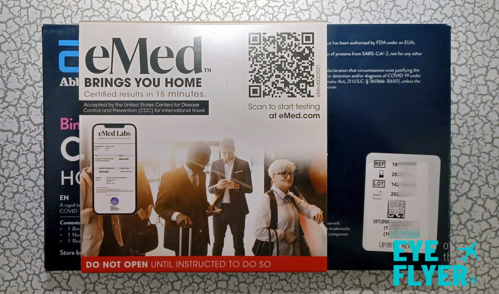 The image shows a box for an eMed COVID-19 home test kit. The packaging highlights that the test provides certified results in 15 minutes and is accepted by the CDC for international travel. There is a QR code for starting the test at eMed.com. The box also features an image of three people, one of whom is holding a phone. A red strip at the bottom reads "DO NOT OPEN UNTIL INSTRUCTED TO DO SO." The background includes text and branding related to the test kit.