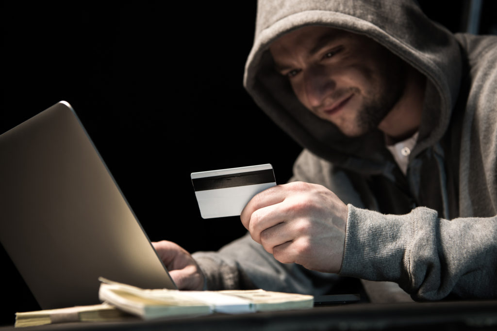 Young hacker in hoodie holding credit card and using laptop