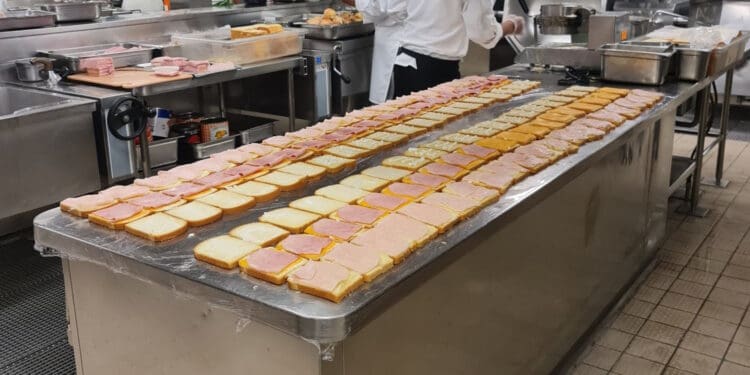 a man in a white coat standing behind a large counter with sandwiches