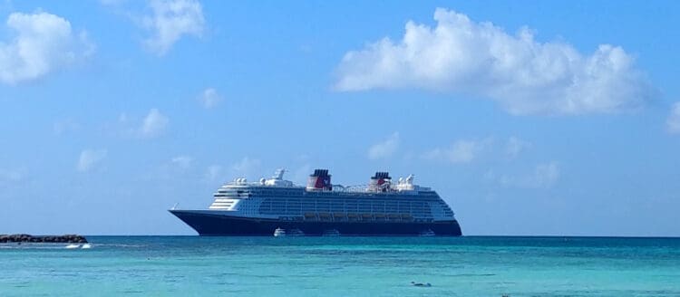 A large cruise ship is sailing on a calm, turquoise sea under a clear blue sky with a few scattered clouds. The ship is positioned in the center of the image, and there is a small rocky outcrop visible on the left side.