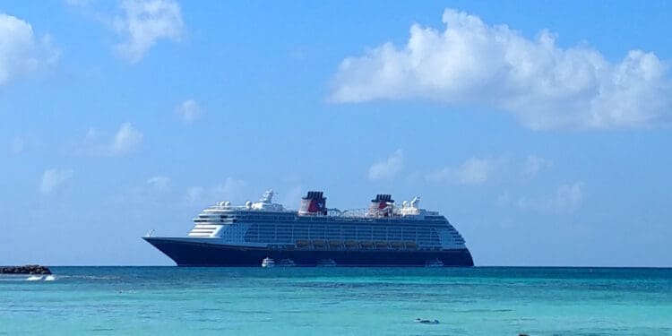 A large cruise ship is sailing on a calm, turquoise sea under a clear blue sky with a few scattered clouds. The ship is positioned in the center of the image, and there is a small rocky outcrop visible on the left side.
