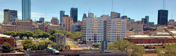 A cityscape featuring a mix of modern and historic buildings under a clear blue sky. In the foreground, there are trees and a few industrial structures. The skyline includes tall skyscrapers and mid-rise buildings, with a prominent glass tower. The scene is bustling with activity, suggesting an urban environment.