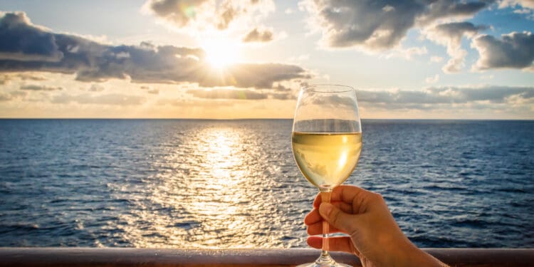 During sunset a female hand toasts the new year with a glass of white wine. Photo taken on a balcony of a cruise ship with a sea view. Cloudy sky.