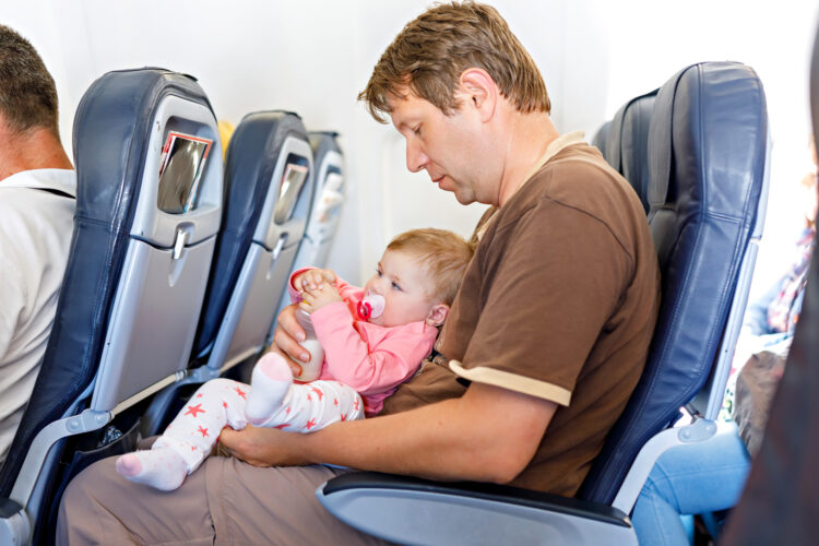 Father holding baby daughter during flight on airplane going on vacations. Baby girl drinking formula milk from bottle. Air travel with baby, child and family concept. Tired man traveling with kids