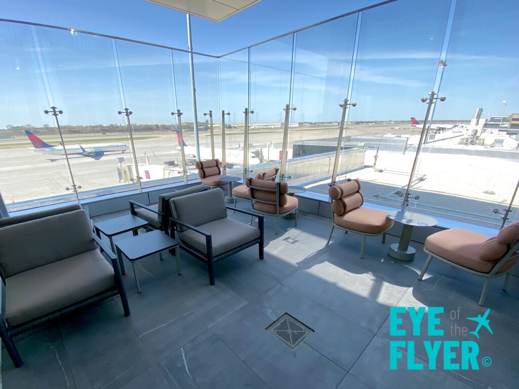 The image shows a modern airport lounge with comfortable seating, including cushioned chairs and small tables. The lounge has large glass windows offering a view of the airport runway, where several airplanes are visible. The sky is clear and blue, and the lounge is well-lit with natural light. The logo "Eye of the Flyer" is visible in the bottom right corner.