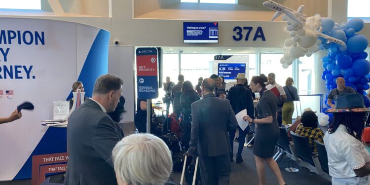 The image shows a busy airport gate area, specifically gate 37A. There are several people standing and walking around, some holding papers or boarding passes. A Delta Airlines sign indicates "Sky Priority" and "General Boarding" areas. In the background, there is a display screen with flight information. A decorative arrangement of blue and white balloons, shaped like an airplane, is visible. The words "Eye of the Flyer" are overlaid on the image.