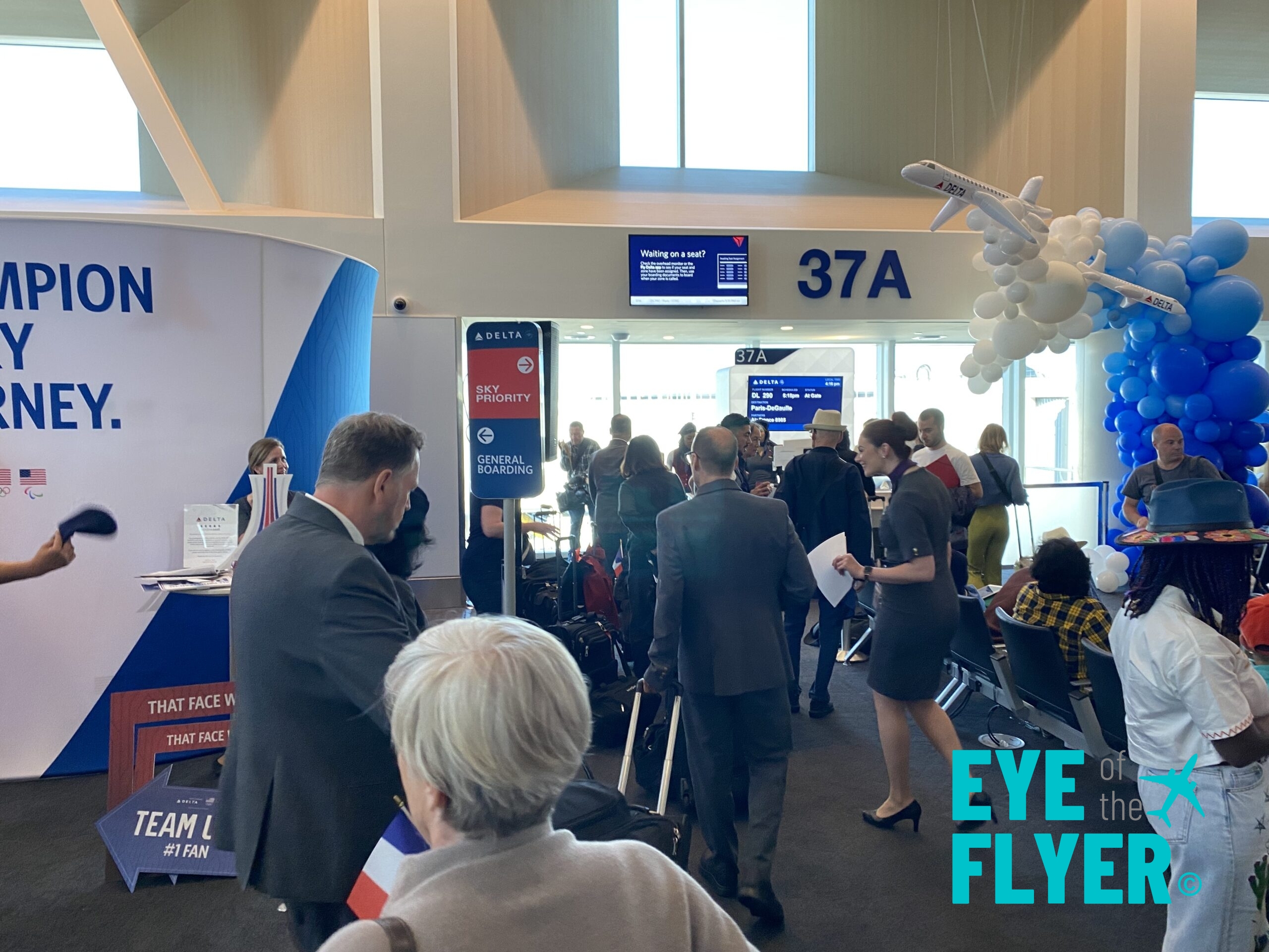 The image shows a busy airport gate area, specifically gate 37A. There are several people standing and walking around, some holding papers or boarding passes. A Delta Airlines sign indicates "Sky Priority" and "General Boarding" areas. In the background, there is a display screen with flight information. A decorative arrangement of blue and white balloons, shaped like an airplane, is visible. The words "Eye of the Flyer" are overlaid on the image.