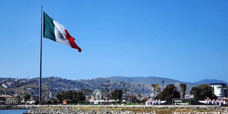 A large Mexican flag is prominently displayed, waving in the wind against a clear blue sky. Below, there is a rocky shoreline and a body of water. In the background, there are hills with scattered buildings and a sign that reads "Puerto de Ensenada." Palm trees and a Ferris wheel are also visible in the distance.