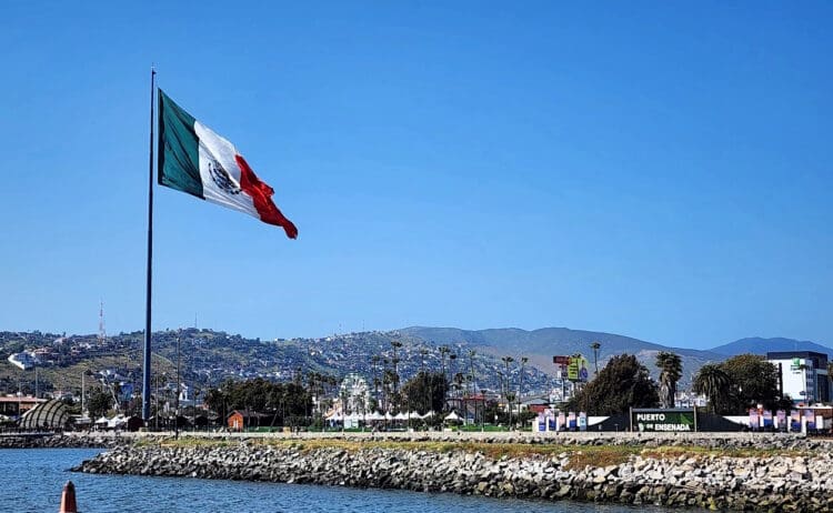 A large Mexican flag is prominently displayed, waving in the wind against a clear blue sky. Below, there is a rocky shoreline and a body of water. In the background, there are hills with scattered buildings and a sign that reads "Puerto de Ensenada." Palm trees and a Ferris wheel are also visible in the distance.
