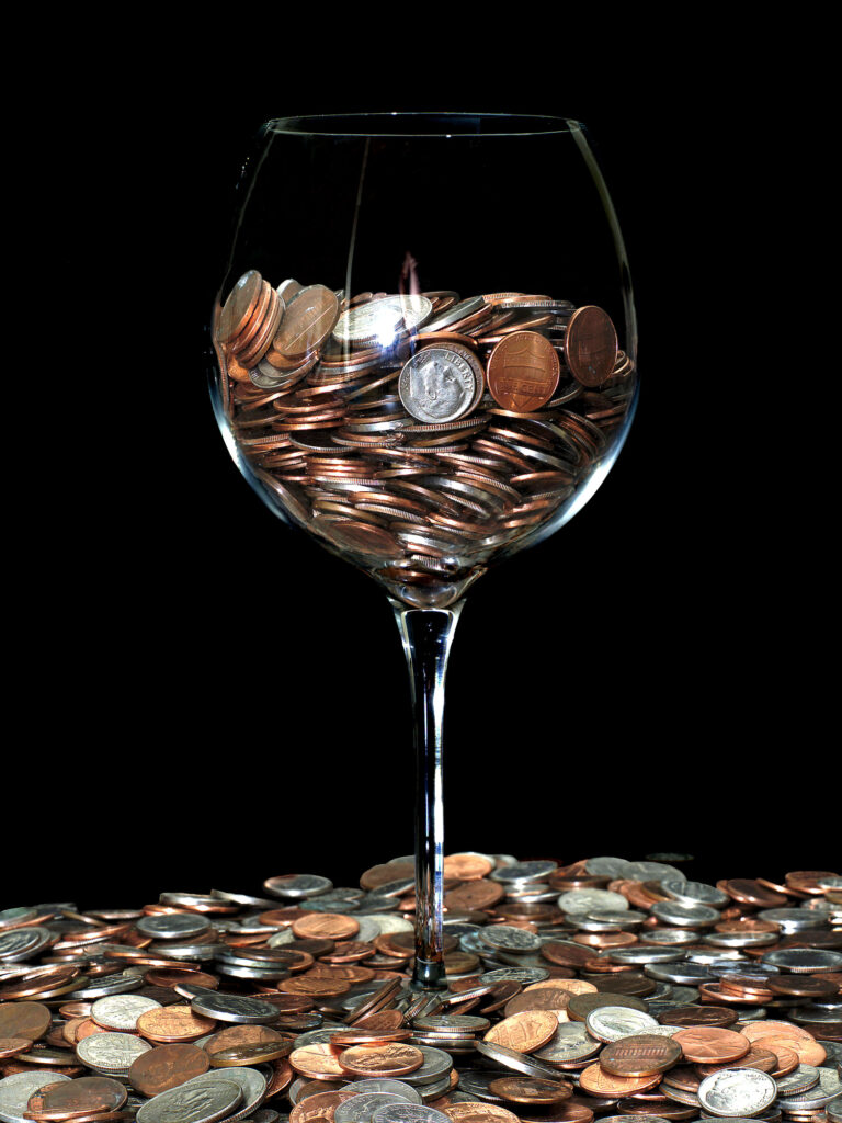 A large wine glass half filled with US coins on a surface covered with US coins.