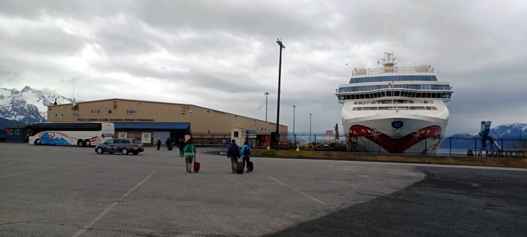 people walking with luggage in a parking lot with a large ship