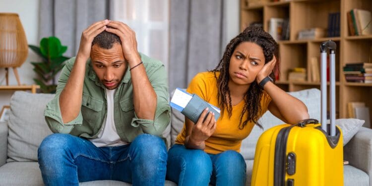 Cancelled flight. Upset, surprised couple after tour cancellation, sitting on sofa at home with travel tickets and suitcase