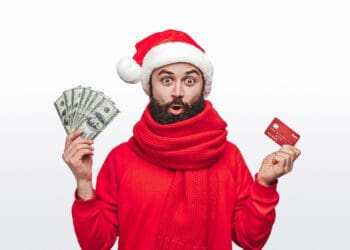 Shocked bearded man in Santa hat holding money and credit card while making Christmas purchases against white background