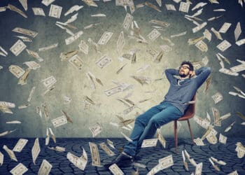 Young, content man sitting on chair in rain of flying bills content with financial independence.