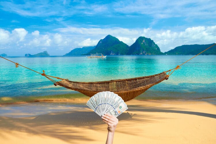 A hammock at the beach with the view of Bacuit Archipelago islands - El Nido, Philippines, with a hand holding up a fan of hundred dollar bills.