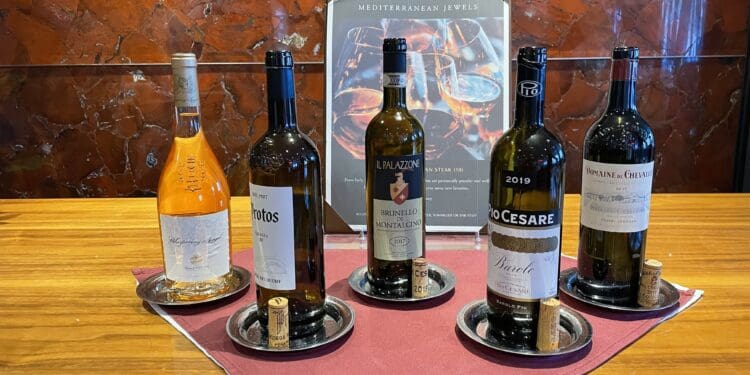The image shows five bottles of wine displayed on a wooden surface. Each bottle is placed on a small round tray with a cork beside it. In the background, there is a sign titled "Wine Tasting Mediterranean Jewels" with an image of wine glasses. The setting appears to be a wine tasting or display area with a rich, textured red-brown wall.