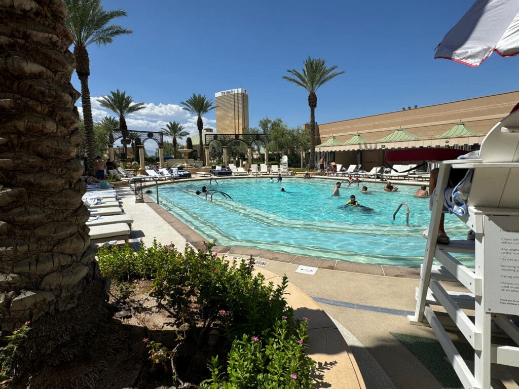 A swimming pool surrounded by lounge chairs and palm trees under a clear blue sky. People are swimming and relaxing in the pool. A lifeguard chair is visible on the right, and a tall building is in the background.