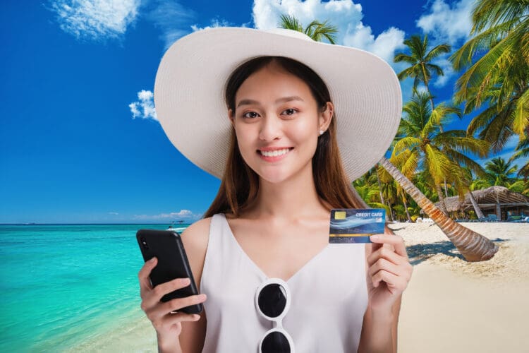 A young woman traveling to an island smiles while posing with her smart phone and credit card