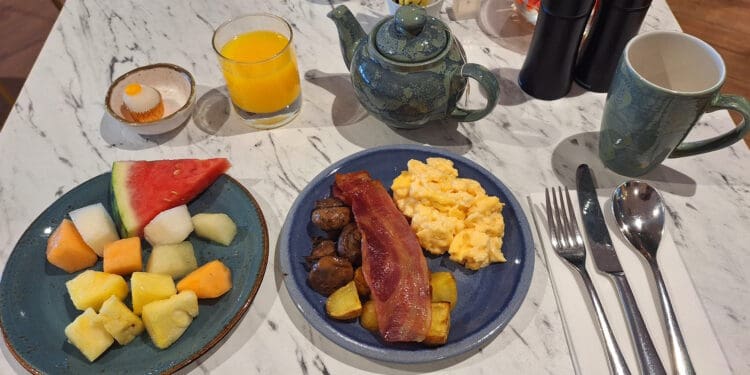 A breakfast setting on a marble table includes a plate of mixed fruit with watermelon, pineapple, and melon pieces, and another plate with scrambled eggs, bacon, mushrooms, and potatoes. There is a small dessert, a glass of orange juice, a teapot, a mug, and cutlery on a napkin.