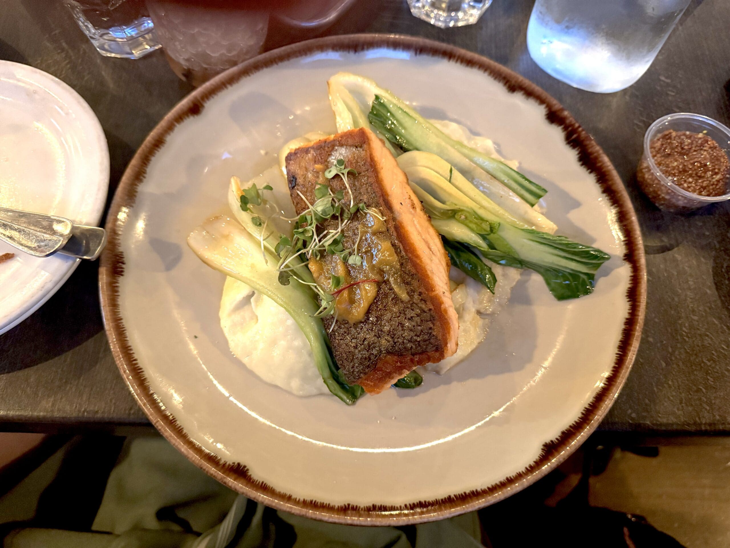 A plate of food featuring a piece of cooked salmon with crispy skin, garnished with microgreens and a sauce. The salmon is served on a bed of creamy mashed potatoes, accompanied by sautéed bok choy. The plate is on a dark table, with a glass of water and a small container of seasoning nearby.