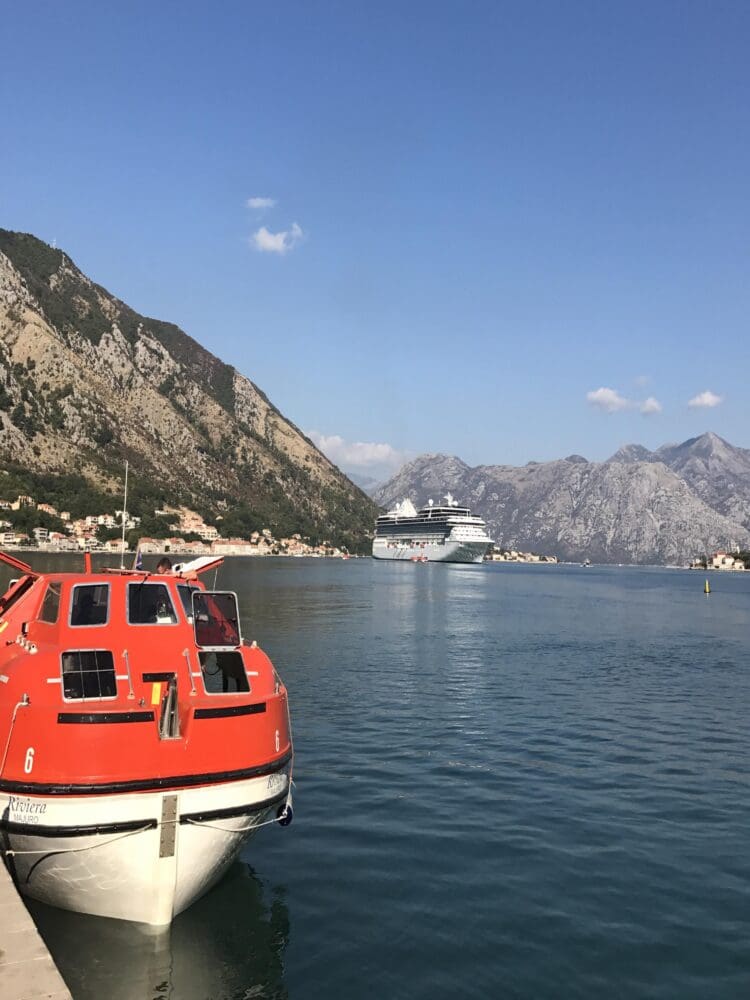 A bright orange lifeboat is docked at a pier in the foreground, with a large cruise ship visible in the distance on a calm body of water. The background features rugged, rocky mountains under a clear blue sky with a few small clouds.