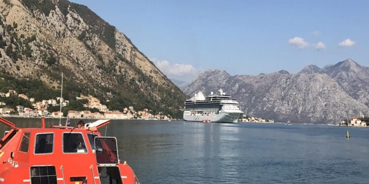 A bright orange lifeboat is docked at a pier in the foreground, with a large cruise ship visible in the distance on a calm body of water. The background features rugged, rocky mountains under a clear blue sky with a few small clouds.