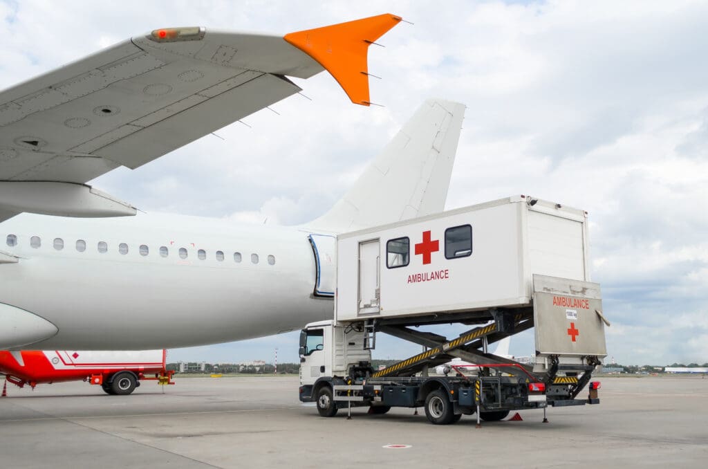 The image shows an airplane on an airport tarmac with a specialized ambulance vehicle positioned next to it. The ambulance is elevated to the height of the airplane's door, suggesting it is used for medical transport directly to and from the aircraft. The ambulance has a red cross and the word "AMBULANCE" on its side. The sky is cloudy in the background.