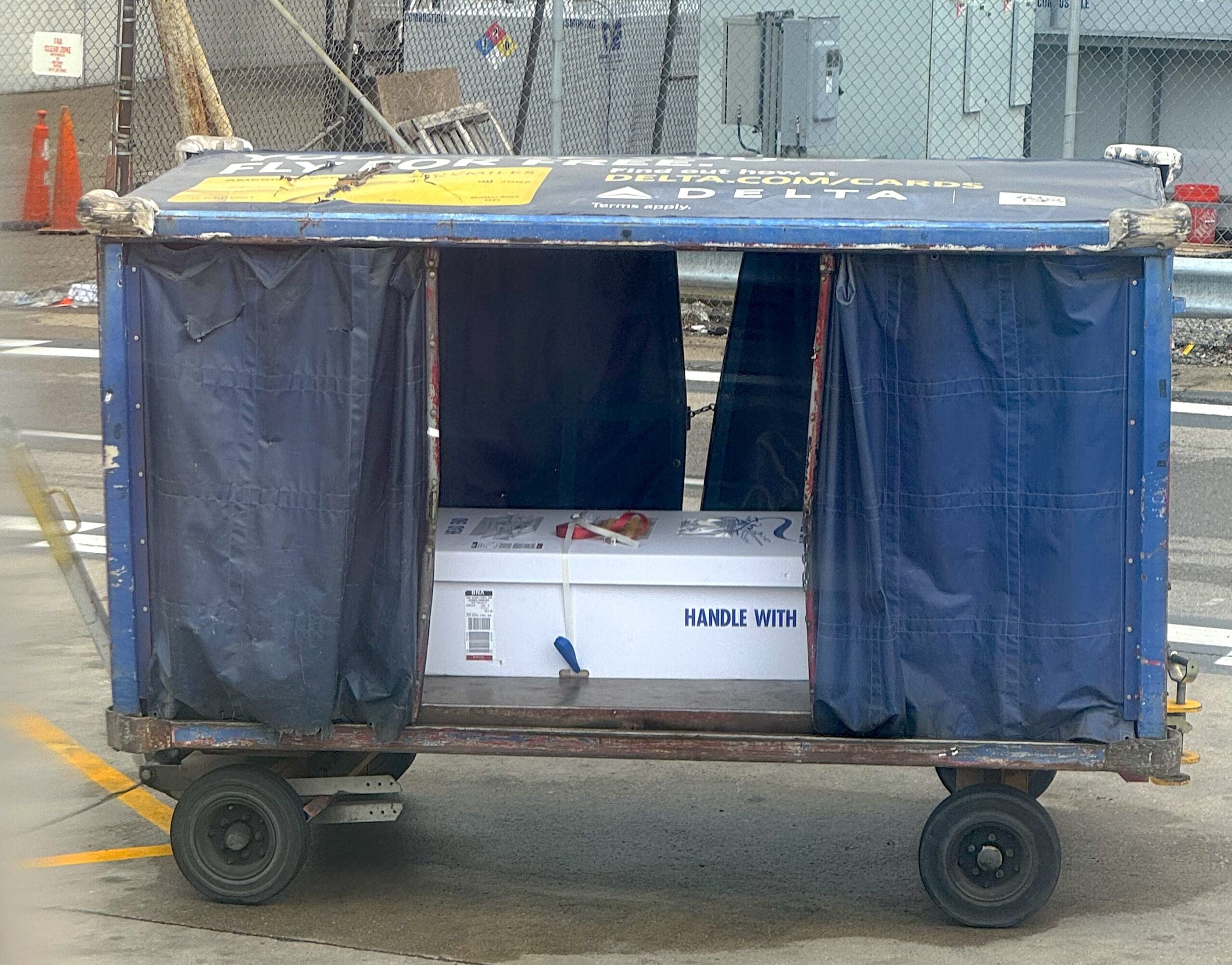 The image shows a blue airport luggage cart with curtains partially open, revealing a large white box inside. The box contains human remains and has "HANDLE WITH CARE" written on it. The cart is on a tarmac area, with a chain-link fence and some orange traffic cones in the background.