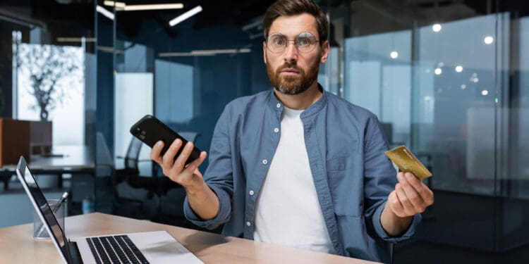 Portrait of disappointed and deceived businessman inside office, mature man looking at camera confused holding bank credit card and phone, employee got rejection error stock photo