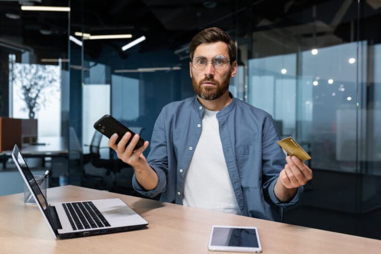 Portrait of disappointed and deceived businessman inside office, mature man looking at camera confused holding bank credit card and phone, employee got rejection error stock photo