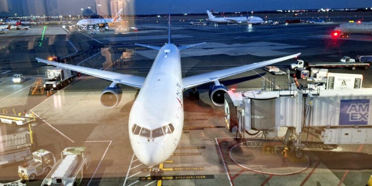 The image shows an airplane parked at an airport gate during twilight. The aircraft is connected to a jet bridge, and various ground service vehicles are nearby, including baggage carts and service trucks. Other airplanes and airport lights are visible in the background on the tarmac.