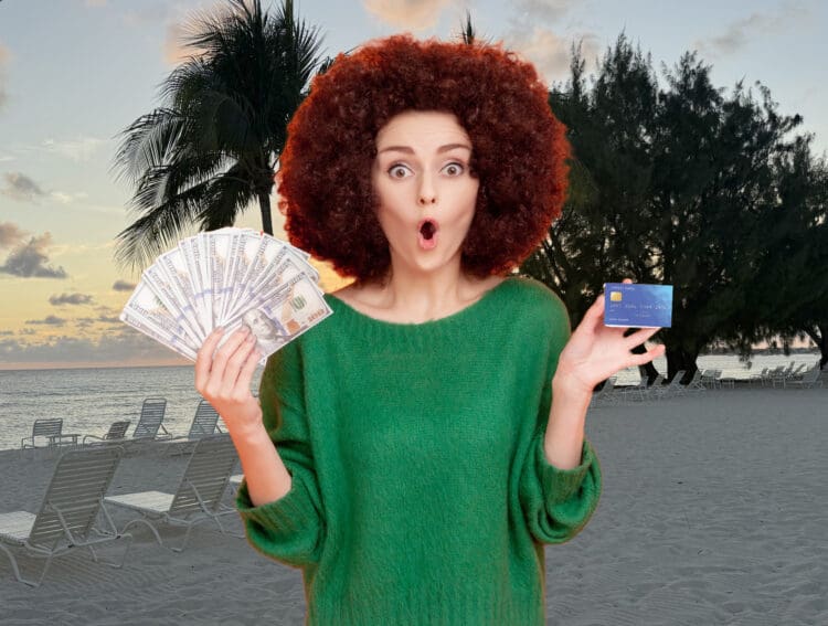 A person with curly red hair is standing on a beach at sunset. They are wearing a green sweater and have an expression of surprise. In one hand, they are holding a fan of hundred-dollar bills, and in the other hand, they are holding a credit card. Palm trees and lounge chairs are visible in the background.