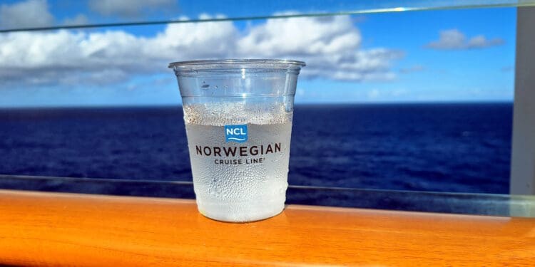 A plastic cup with the "Norwegian Cruise Line" logo is placed on a wooden railing. The cup contains a clear liquid with condensation on the outside. In the background, there is a view of the ocean and a partly cloudy sky.
