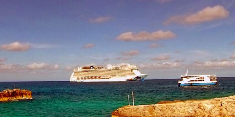 A large cruise ship is sailing on a calm ocean under a partly cloudy sky. In the foreground, a smaller boat is visible near rocky outcrops. The water is a deep blue-green, and the scene conveys a sense of tranquility and travel.