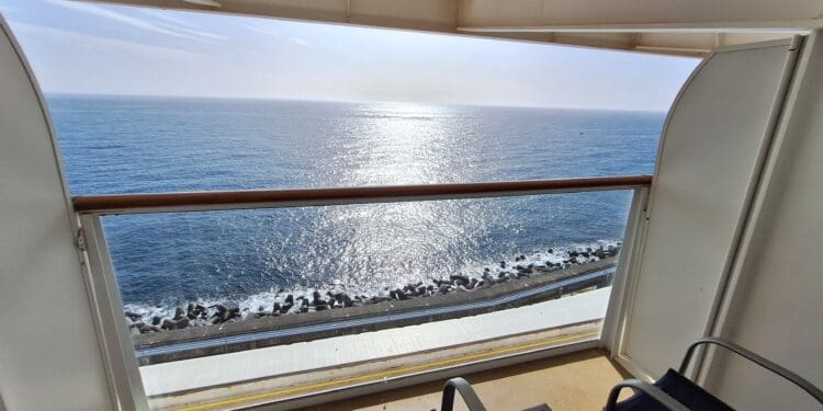 A view from a balcony overlooking the ocean. The sun is reflecting off the water, creating a shimmering effect. The balcony has a glass railing with a wooden handrail, and there are rocks and a breakwater visible along the shoreline below. A chair is partially visible in the foreground.