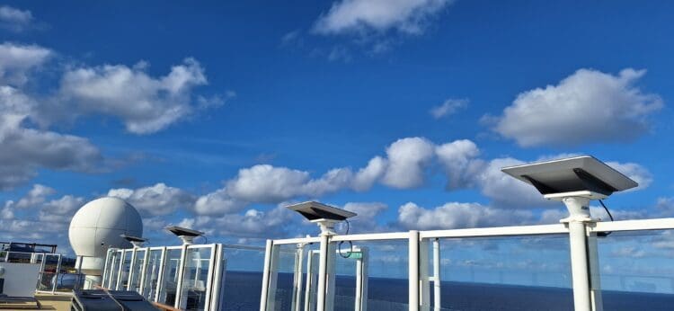 The image shows a clear blue sky with scattered white clouds. In the foreground, there are several satellite dishes mounted on a structure, possibly on a ship or a building, with a large white spherical radar dome visible on the left. The ocean is visible in the background.