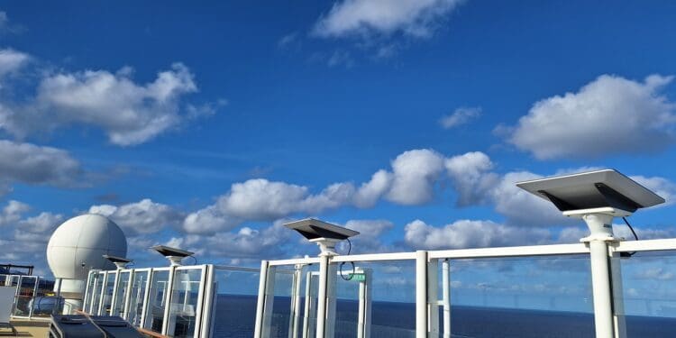 The image shows a clear blue sky with scattered white clouds. In the foreground, there are several satellite dishes mounted on a structure, possibly on a ship or a building, with a large white spherical radar dome visible on the left. The ocean is visible in the background.