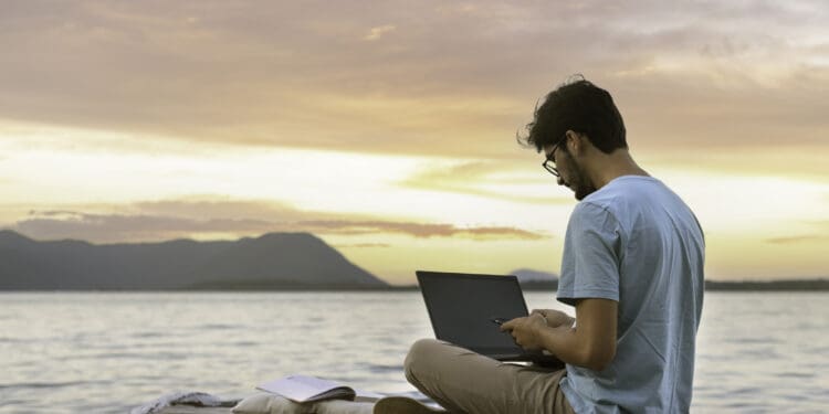 young digital nomad man sitting on wooden pier at sea working on internet remotely at sunset - Traveling with a computer - Online dream job concept